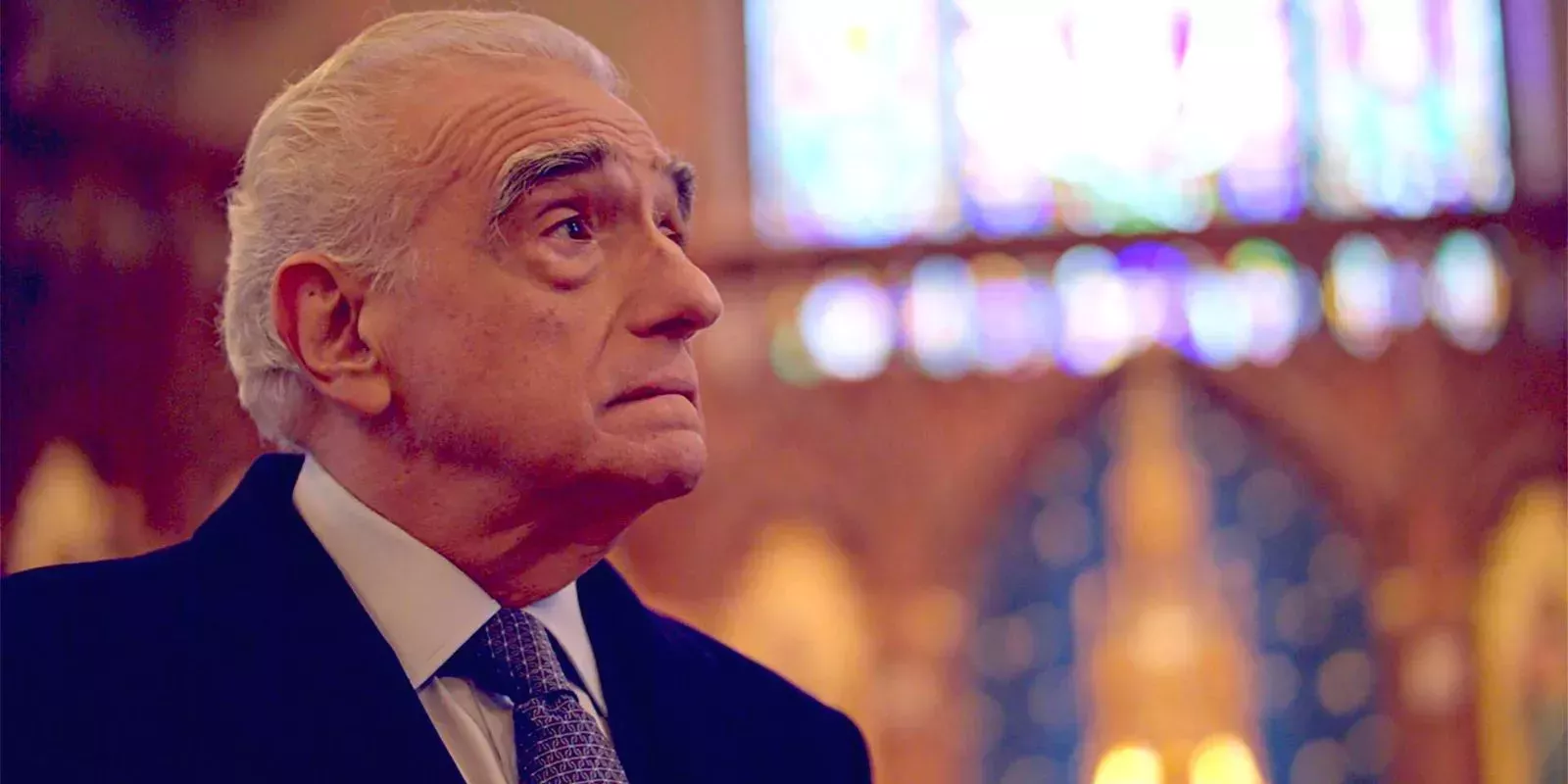 Martin Scorsese looking pensive in a blurry cathedral dressed in suit and tie