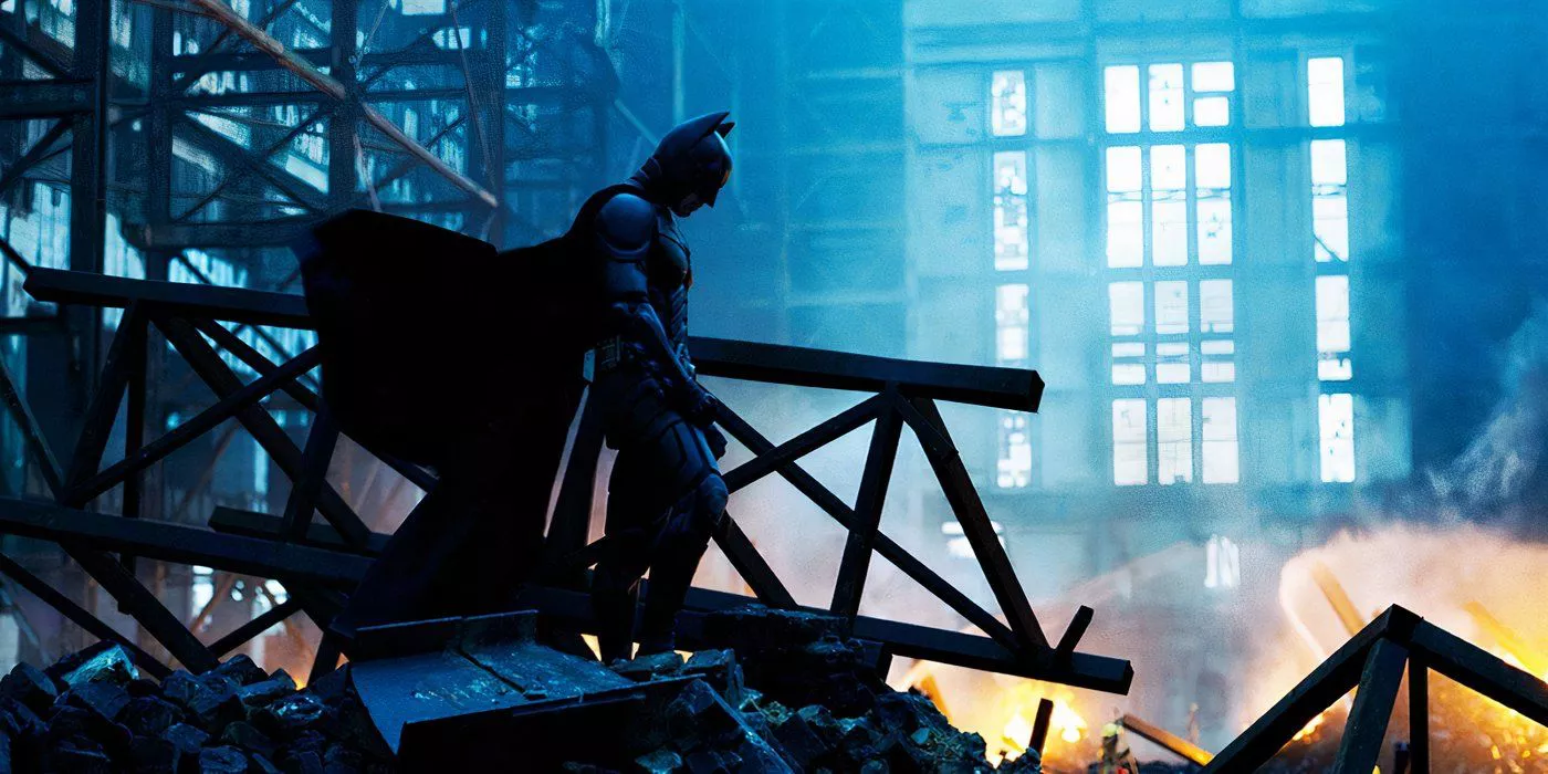 Bruce Wayne/Batman stand atop a pile of rubble from the building that blew up and killed Rachel in The Dark Knight.